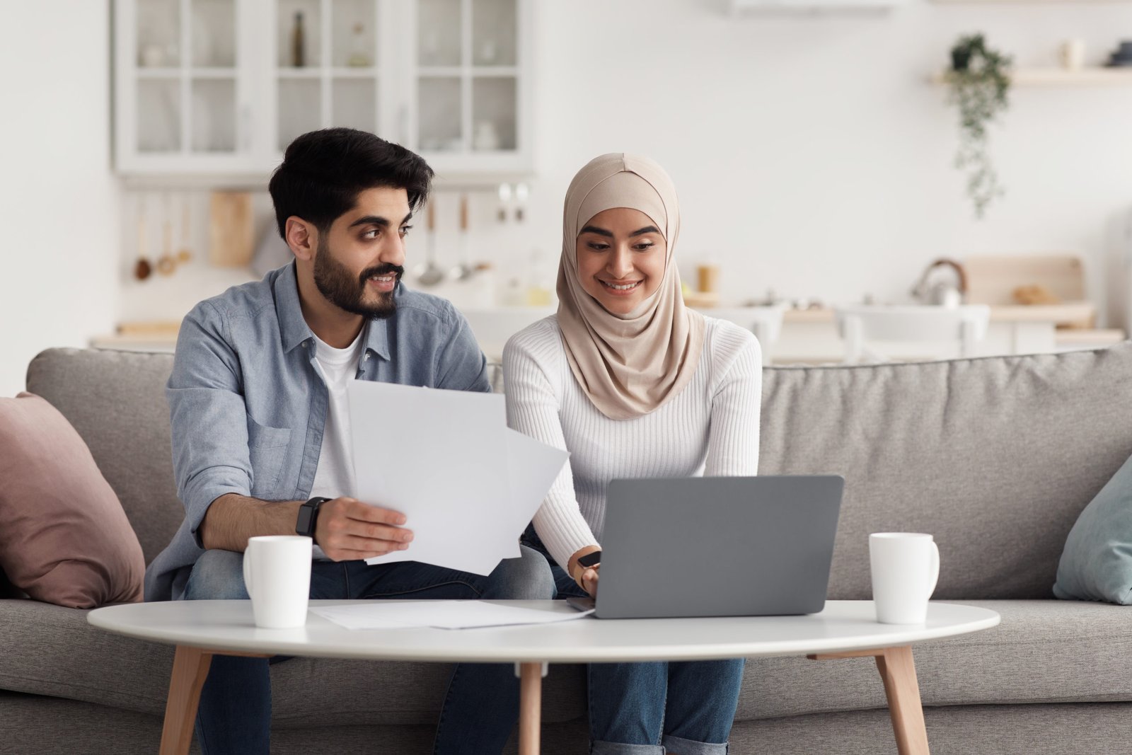 Bills, domestic taxes, home bookkeeping, paperwork together. Smiling millennial muslim female in hijab and male drinking coffee and paying for service on laptop in living room interior, free space